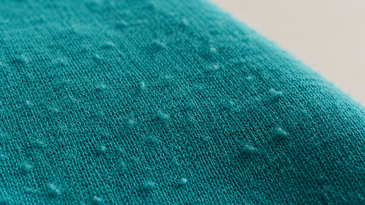 Close-up of a teal cotton knit surface showing fuzz and small pills catching the light.